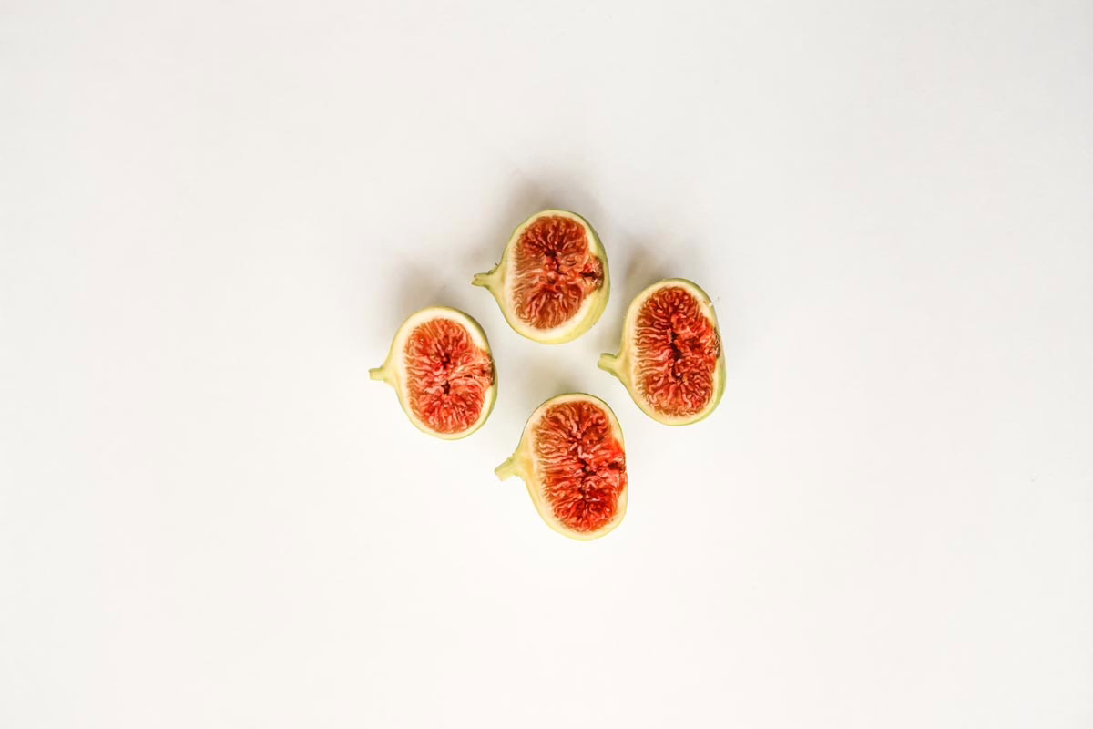 Four halved figs, their red pulps facing up, arranged in a square on a white surface. The figs have light green skins and are ripe and juicy.