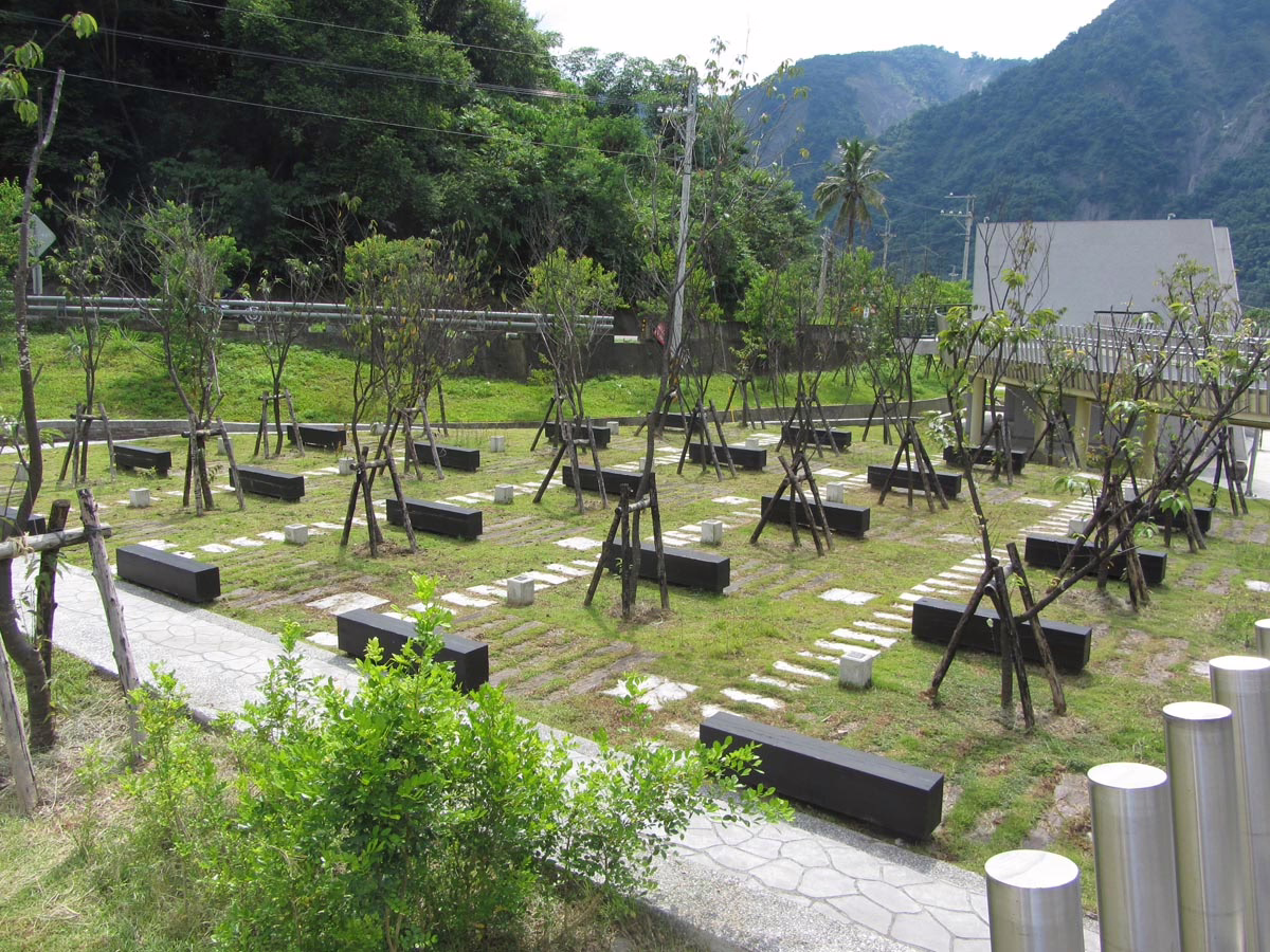 Serene park scene with benches arranged among young trees supported by stakes. A stone pathway meanders through the green space, with mountains visible in the background.